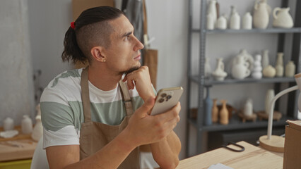 Fototapeta premium Man potter in apron holding smartphone and resting chin on fist while seated at wooden table surrounded by ceramics and shelves in studio; quiet contemplation.