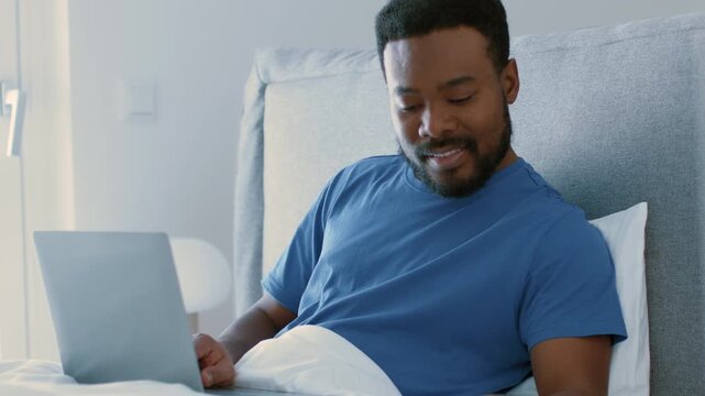 A man with dark skin sits comfortably in bed, working on a laptop. The setting is bright and modern, with light coming through the window. He appears focused and engaged in his tasks.