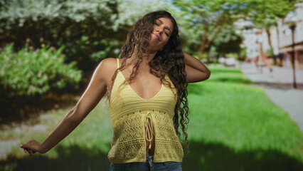 Fototapeta premium Young woman with hand to hair and arm out, wearing yellow knit top showing midriff on street; carefree joy.