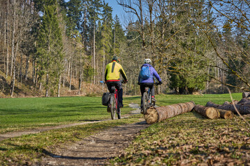 Fototapeta premium Happy senior couple enjoying an active lifestyle, riding electric bikes on a forest trail in spring. Outdoor recreation, healthy aging, and leisure time in nature on a sunny day.