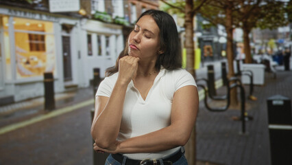 Fototapeta premium Woman with hand on chin, eyes closed and crossed arms on city street near trees and bollards, wearing white top and jeans; contemplation uncertainty.