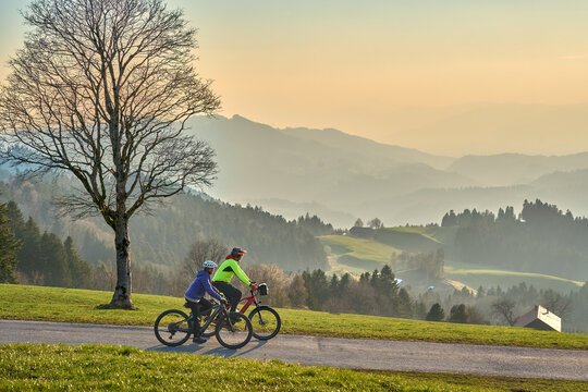 Active senior couple riding electric bikes through scenic Bregenzerwald in Austria during golden evening light. Enjoying nature, mountains, and healthy outdoor lifestyle in spring countryside.
