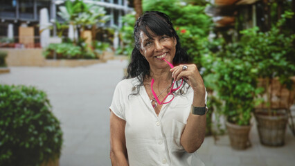 Fototapeta premium Woman senior hispanic holding pink glasses to mouth while smiling and tilting head in building atrium; playful confidence.