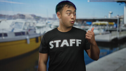 Fototapeta premium Man in staff t shirt on boat gives thumbs up with hand beside building in bright sunlight; approval.