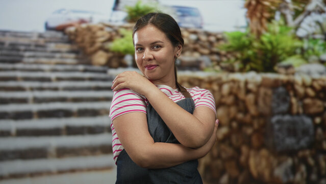 Woman hugs her arms across chest wearing striped shirt and apron on street near stone steps and plants; confidence.