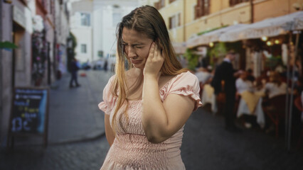 Fototapeta premium Woman holding her ear with one hand and wincing in visible pain at a busy street cafe with tables and diners; earache distress.