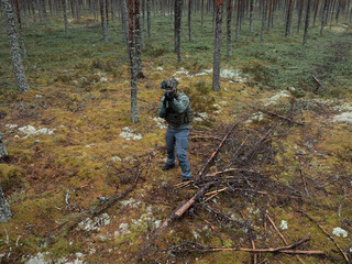 Obraz premium Full length view from a drone of a modern soldier in tactical gear aiming a rifle at a quadcopter in a dense pine forest.