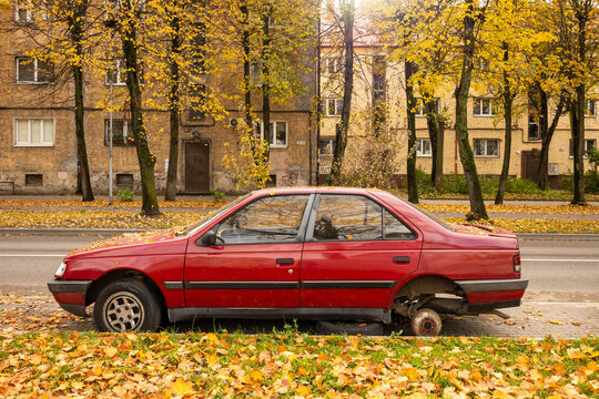 Damaged red car with missing rear wheel parked on street with autumn trees and fallen leaves