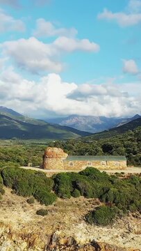 Historic stone building surrounded by lush greenery and mountains under a bright blue sky in the Delta du Fango, Corsica. Scenic natural landscape with dramatic clouds and rugged terrain.