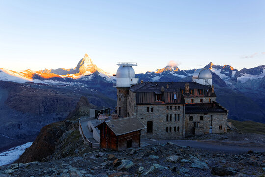 Sunrise scenery of Matterhorn among impressive alpine mountains with golden sunlight on the summit and the famous Gornergrat Observatory on a rocky ridge in foreground in Zermatt, Valais, Switzerland