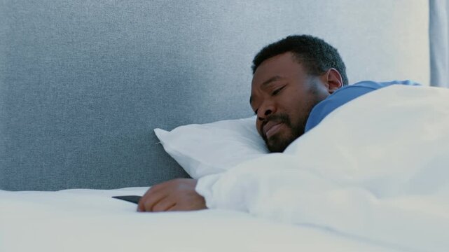 A man with dark skin is sleeping in a cozy bedroom. The setting includes a neatly made bed with white sheets. A smartphone rests on the bedside table, indicating the time of day.