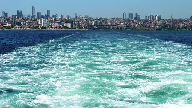 Wide shot from the stern of a fast-moving passenger ferry showing turbulent wake and white foam trail on turquoise sea, with the distant Istanbul skyline visible along the coastline near Bostanci