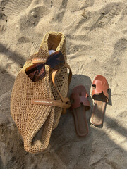 A stylish woven straw tote bag with a eyeglasses, paired with brown leather sandals on sandy beach ground, evoking summer vibes.