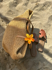 A stylish woven straw tote bag with a vibrant orange flower barrette, paired with brown leather sandals on sandy beach ground, evoking summer vibes.