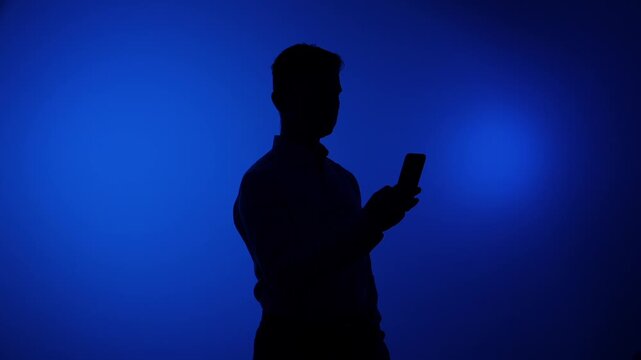 Male profile silhouette standing in front of a vibrant blue backlit background, actively using a mobile phone, representing digital communication, social media, and technology connection