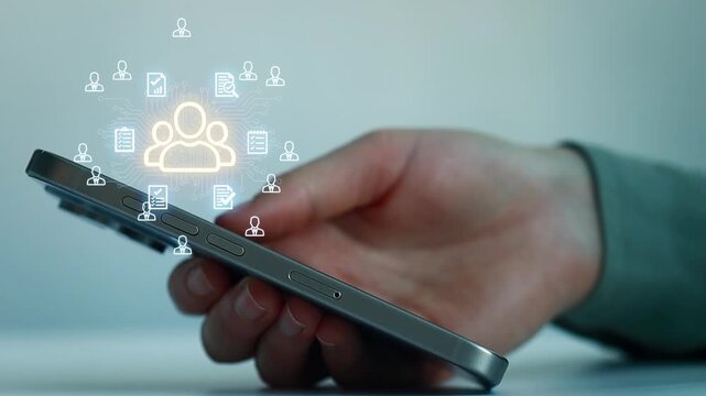 Hand holds smartphone with icons representing communication and data exchange on screen in a modern workspace setting.