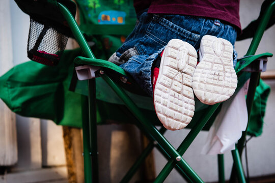 Close-up of a young child's legs in blue jeans and white canvas sneakers dangling from a green folding camping chair, a classic image of outdoor summer childhood.