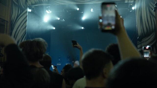 Dynamic concert scene from audience perspective. Crowd records live show with phones, surrounded by beaming stage lights and smoke effects, capturing an immersive entertainment experience