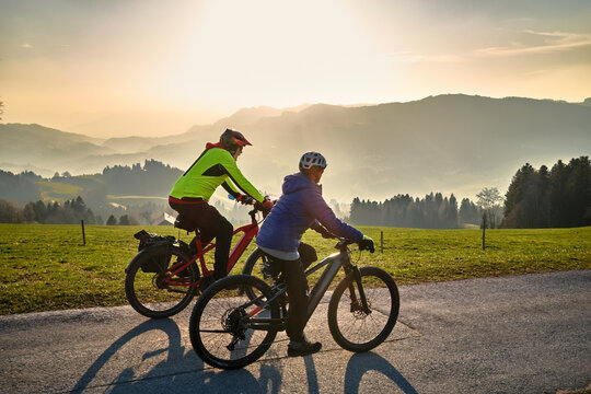 Active senior couple riding electric bikes through scenic Bregenzerwald in Austria during golden evening light. Enjoying nature, mountains, and healthy outdoor lifestyle in spring countryside.
