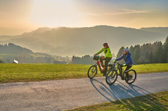 Active senior couple riding electric bikes through scenic Bregenzerwald in Austria during golden evening light. Enjoying nature, mountains, and healthy outdoor lifestyle in spring countryside.
