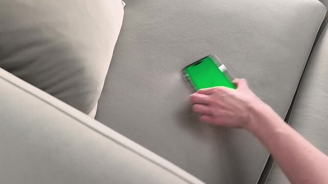 Smartphone with green screen lies on sofa and female hand picks it up at home. Device with chroma display is ready for interaction. Concept app advertising, mockup template, mobile usage,