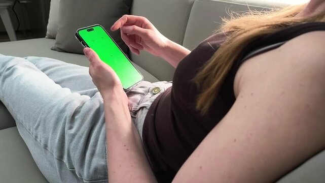 Woman uses smartphone with green screen while sitting on sofa from over shoulder view. Female scrolls mobile interface with chroma display at home. Concept app preview, mockup template,