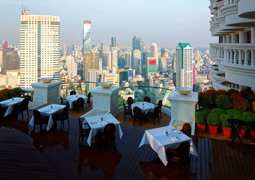 View from a rooftop restaurant in Bangkok, the fast developing capital city of Thailand, with tables on the terrace in foreground & MahaNakhon Tower amid skyscrapers in background under blue sunny sky
