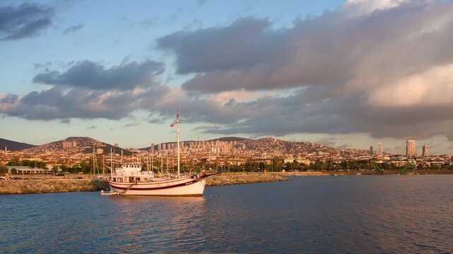 White wooden fishing boat with tall masts moored at the coast of Kucukyali, Maltepe, Istanbul, with residential buildings and Marmara Sea coastline in the background at sunset