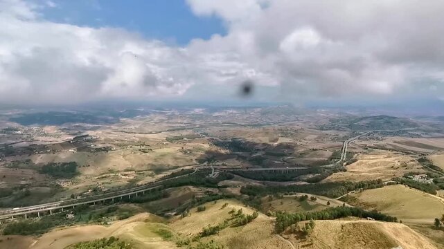Scenic aerial view of rolling hills and valleys in the Province of Enna, Sicily, showcasing a winding road and lush greenery under a cloudy sky