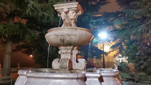 Proserpina Fountain in Enna, Sicily, illuminated at night, showcasing intricate stonework and flowing water, surrounded by lush greenery and atmospheric lighting