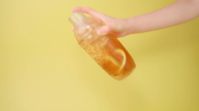 Hand shaking clear shaker bottle making refreshing iced smashed lemon tea with green lime slices and ice cubes on bright yellow background
