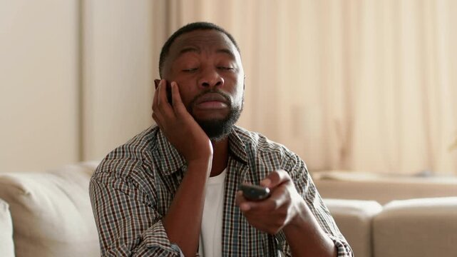 A man with dark skin sits on a sofa in a well-lit living room. He looks directly at the camera while holding a remote control, engaged in watching television.