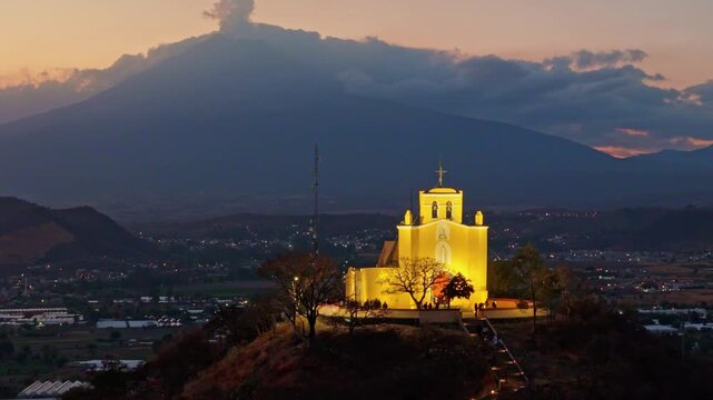Illuminated hilltop church at dusk with Popocatepetl volcano eruption cloud in Atlixco, Mexico