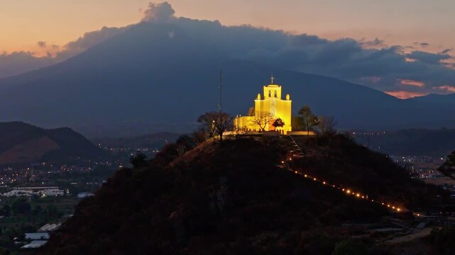 High aerial drone view of hilltop church surrounded by city of Atlixco at sunset, Puebla, Mexico