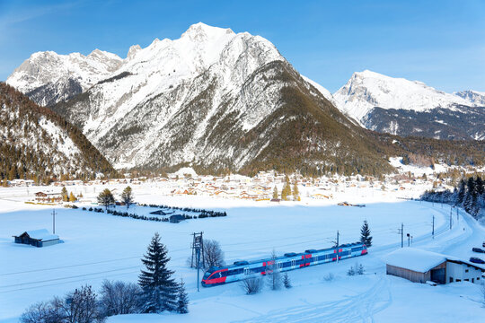 A regional train traveling thru a railway curve in a valley covered by heavy snow & the Scharnitz village lying at the foot of majestic Alpine mountains on a brisk sunny winter day, in Tyrol, Austria