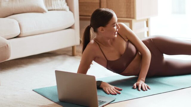 A woman is exercising on a mat at home while following a workout on her laptop. She is engaged in a fitness routine in a cozy living room setting with natural light.