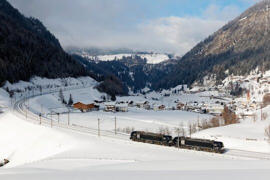 Two connected locomotives traveling thru a railway curve in a valley covered by heavy snow & St. Jodok am Brenner village lying under majestic Alpine mountains on a sunny winter day, in Tyrol, Austria