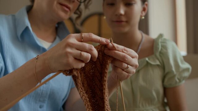 Focus on hands of mature grandmother instructing young granddaughter using needles and yarn while knitting together at home