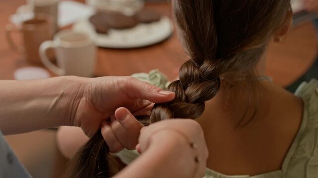 Close up shot of hands of mother plaiting hair of her daughter while bonding at home