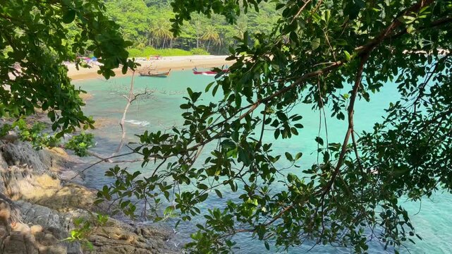 Secluded tropical beach with turquoise water and longtail boats, viewed through dense green foliage along a rocky shoreline in Phuket, Thailand