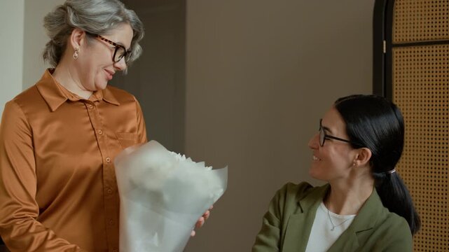 Tracking shot of mature Caucasian mother giving bouquet of white flowers to adult daughter as present in living room