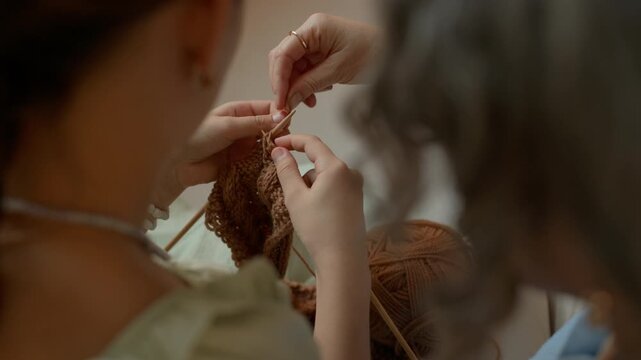 Over the shoulder view of young girl sitting next to grandmother instructing her while knitting together using needles and yarn