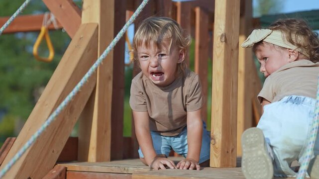 Upset child crying on playground. Emotional toddler expressing frustration outdoors. Child feeling upset while playing. Kids on wooden playground. Crying child in summer light. Child upset playground.