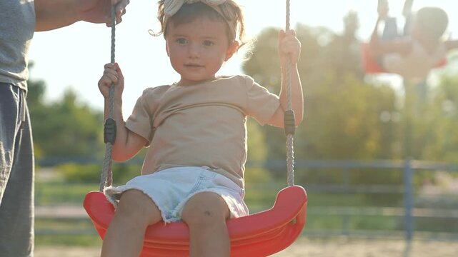 Happy Father pushing child on swing in playground. Happy child on swing enjoying playful moment with father outdoors. Happy Family bonding at playground. Child laughs swinging high on playground swing