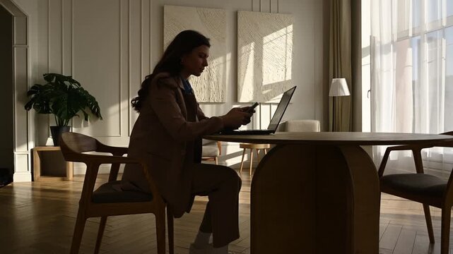 Professional woman in a suit seated at a round table in a sunlit modern office, focused on her smartphone with a laptop nearby as morning light casts her silhouette through a large window