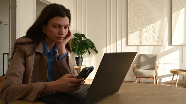 Young businesswoman working on her laptop computer, getting distracted by her smartphone and starting to scroll, browsing social media instead of working, procrastinating at her workplace