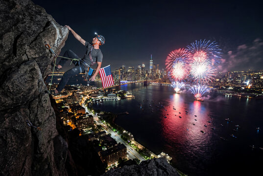 Brave rock climber with American flag climbing steep cliff at night watching Independence Day fireworks over a city skyline