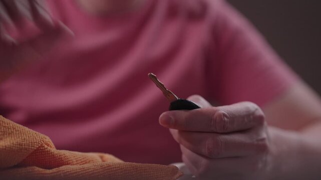 Close-up hand of person disinfecting modern car key with sanitizing spray and cleaning it with orange microfiber cloth for virus and germ prevention sitting at table. Shooting in slow motion.