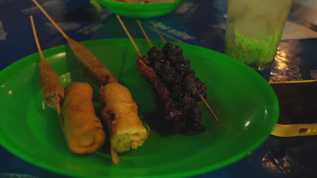 A close-up shot captures a person selecting a snack from an assortment of traditional Indonesian fried appetizers and meat satay served on a green plate at a food stall.