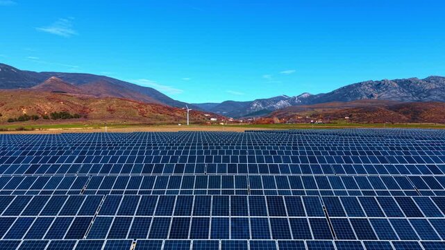 Footage over the multiple modern solar panels. Approaching the wind turbine at the mountain foot in the backdrop.